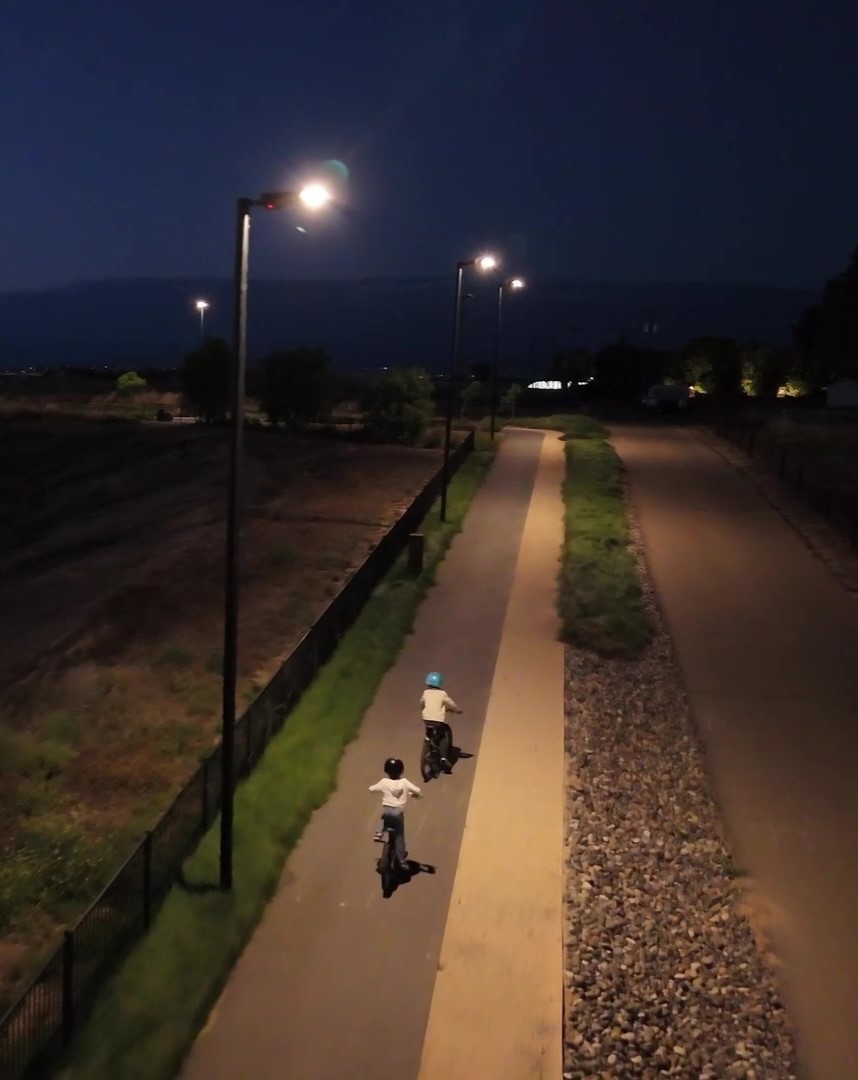 Two children biking along a lit paved path at night, with tall streetlights and a grassy area to the side.